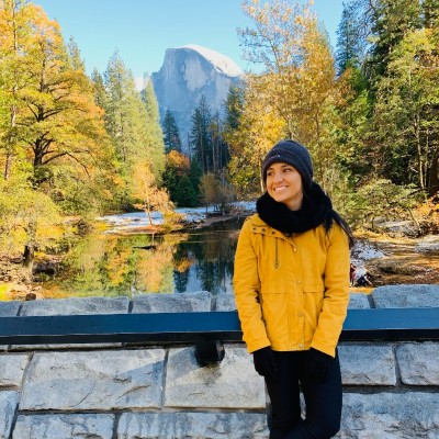 Cariny smiling in a yellow jacket in Yosemite Valley, with Half Dome and fall trees behind her